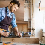 Un menuisier à Seyssel en chemise à carreaux utilise un rabot à main sur un bloc de bois brut, dans un atelier lumineux