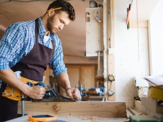 Un menuisier à Seyssel en chemise à carreaux utilise un rabot à main sur un bloc de bois brut, dans un atelier lumineux