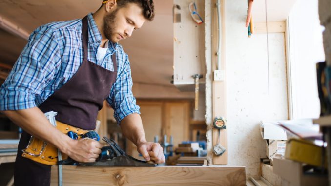 Un menuisier à Seyssel en chemise à carreaux utilise un rabot à main sur un bloc de bois brut, dans un atelier lumineux
